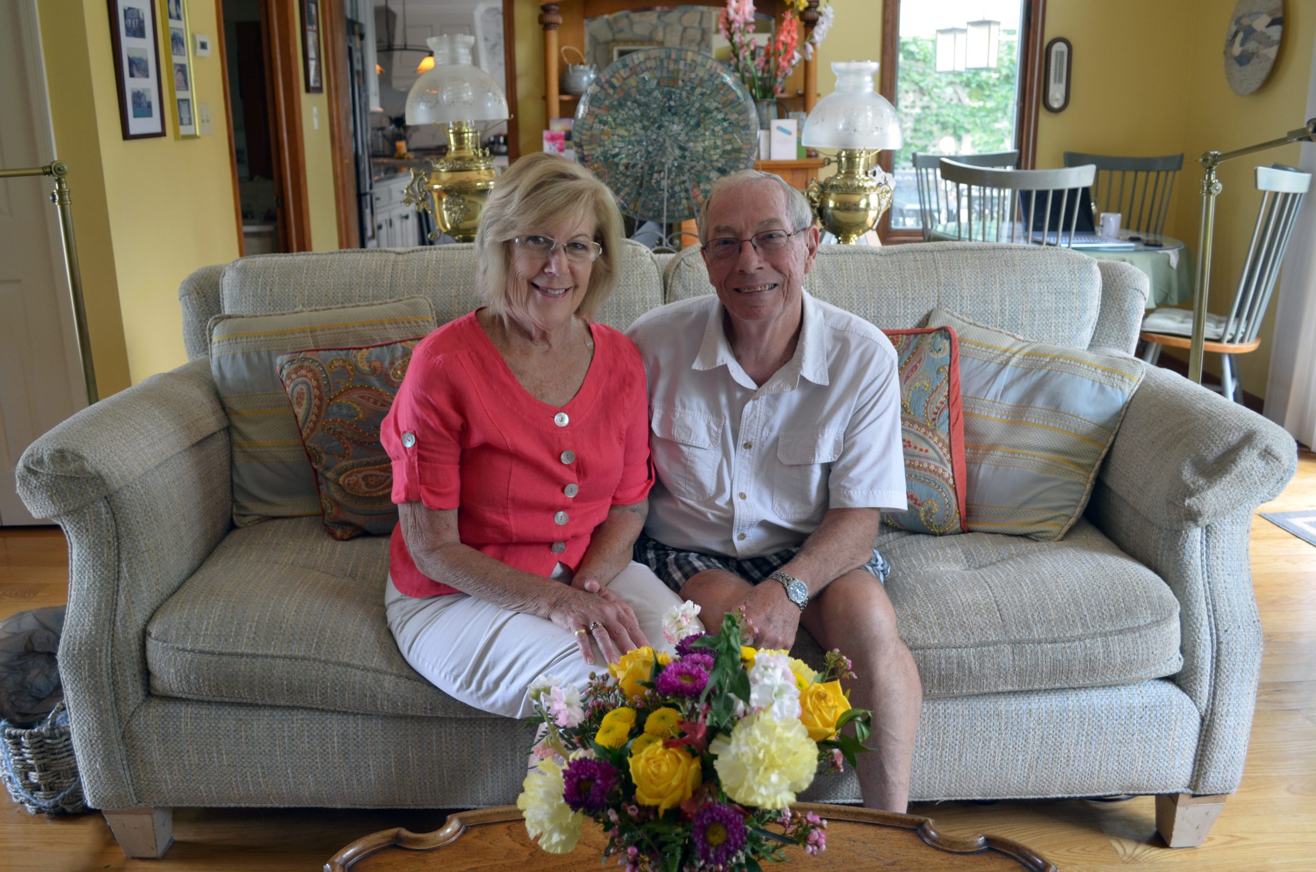 Elderly owners of Central NY bed and breakfast seated on sofa and smiling with flowers