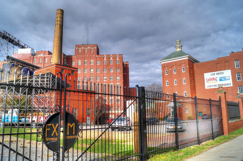 Front view of Central NY brewing company building with red brick fascia and green fencing on a cloudy day.