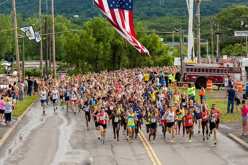 Aerial front view of large running race with American flag, fire engine, and foliage in the background.