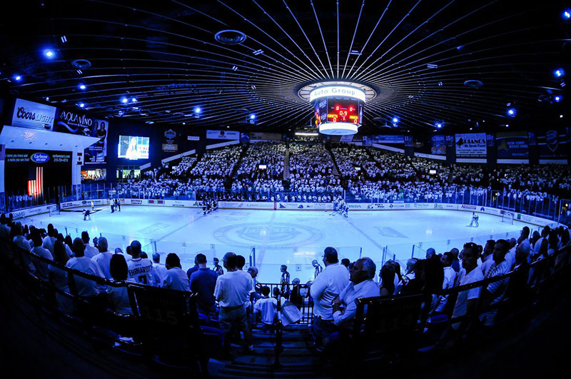 Rinkside view of Central NY hockey arena with blue lighting and spectators.